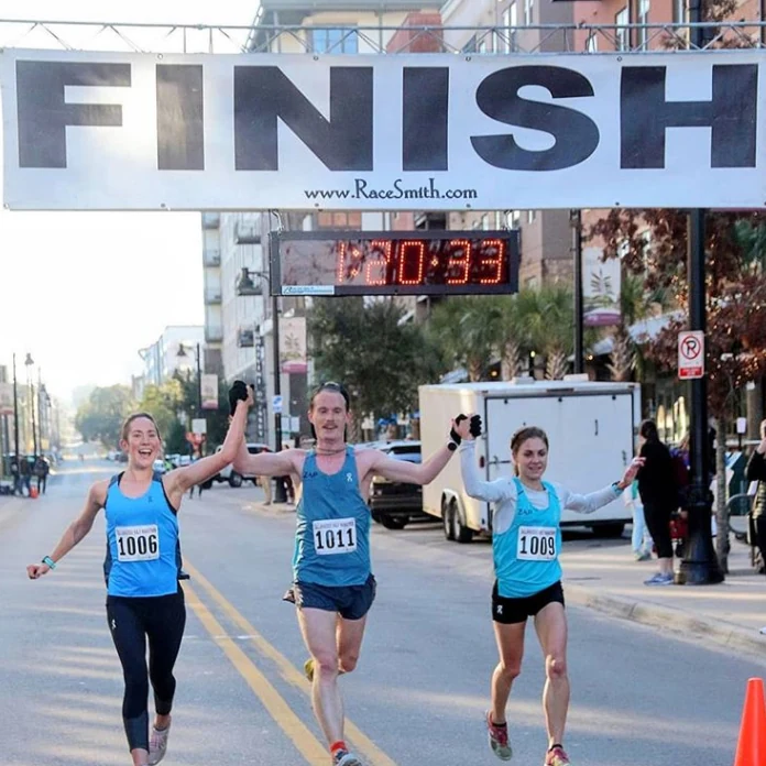 Three runners crossing the finish line side by side with hands raised, race clock reading 1:20:33
