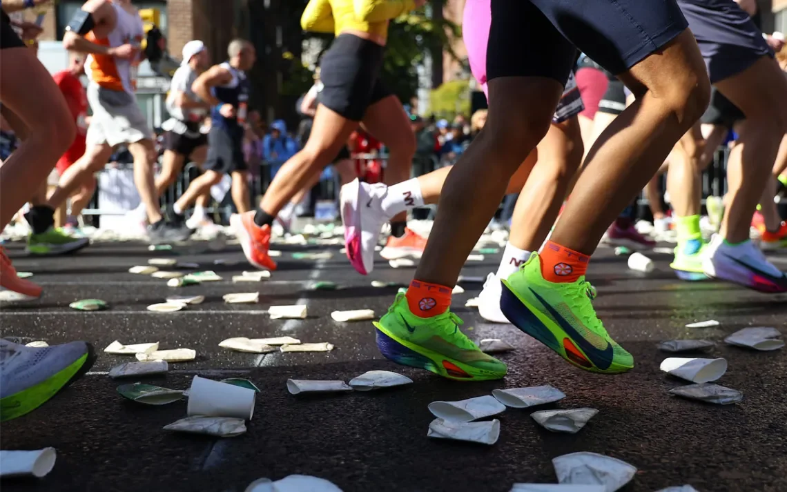 A runner between two aid-station tables refilling a paper water cup mid 10K, hands on the table edge.