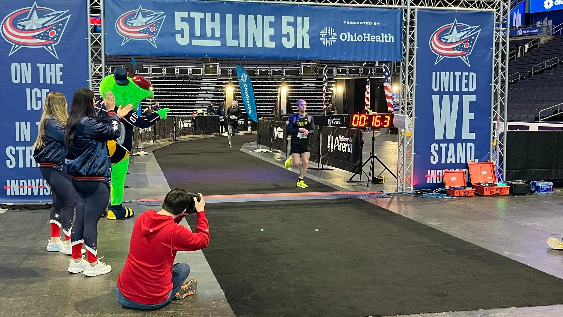 A runner hands on knees catching their breath after a 5K finish, digital race clock blurred behind.
