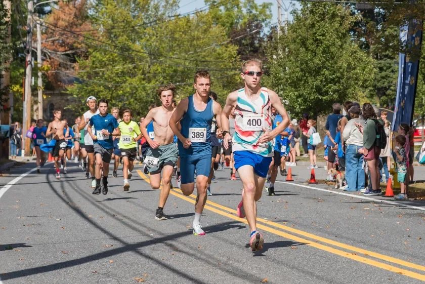 Close-up of a runner's wrist watch flashing a kilometer split, sweat on the forearm, sunlit park path blurred behind.