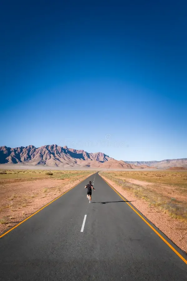 A solo runner as a small distant figure on an empty two-lane road at golden hour, long shadows across the pavement.