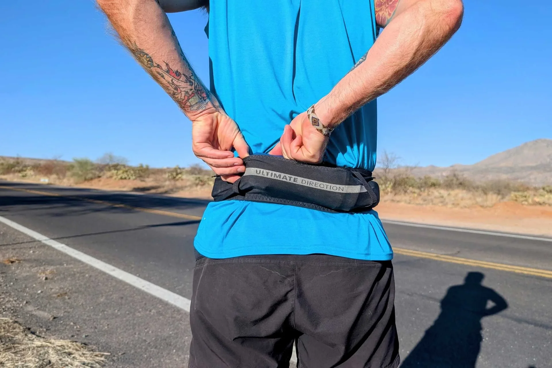 A runner's waist and hands tucking an energy gel into a race belt on a quiet road at sunrise.