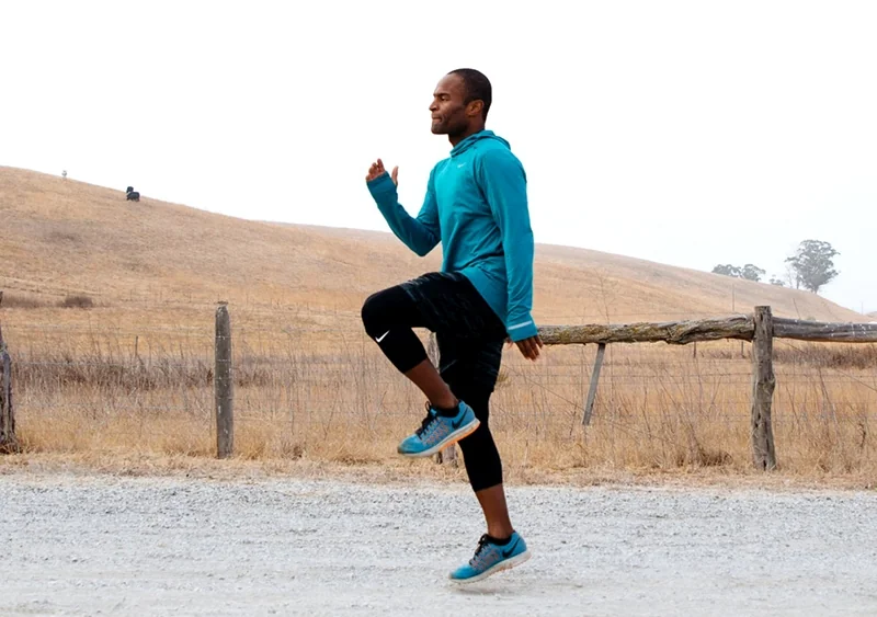 A runner performing a tall standing marching drill on grass with the lead knee lifted to hip height.