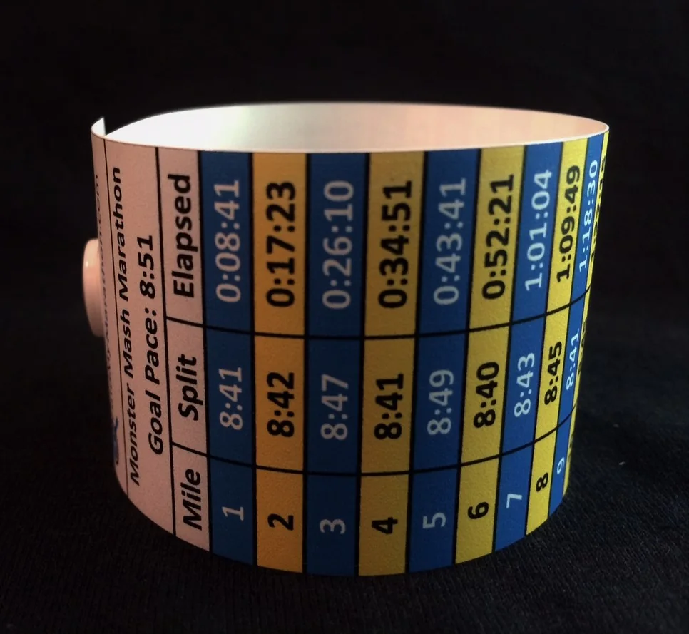A weathered race-day pace band laid across an open marathon course map with a stopwatch and pen beside it.