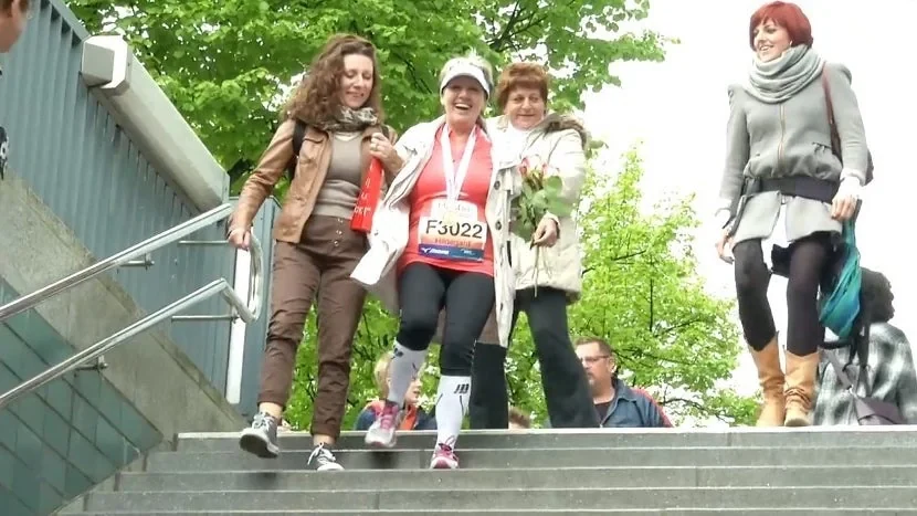 A marathon finisher in her bib and medal laughs as two friends help her down a flight of stairs.