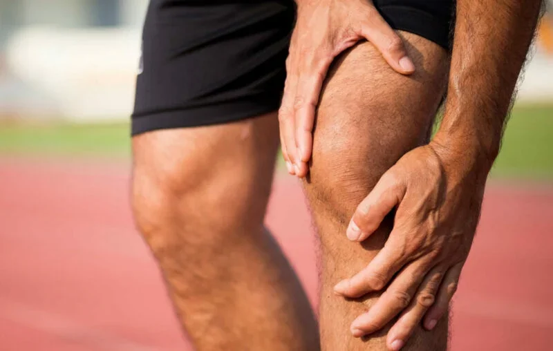 A runner pausing at the side of a sunlit gravel path, one hand resting lightly above the kneecap.