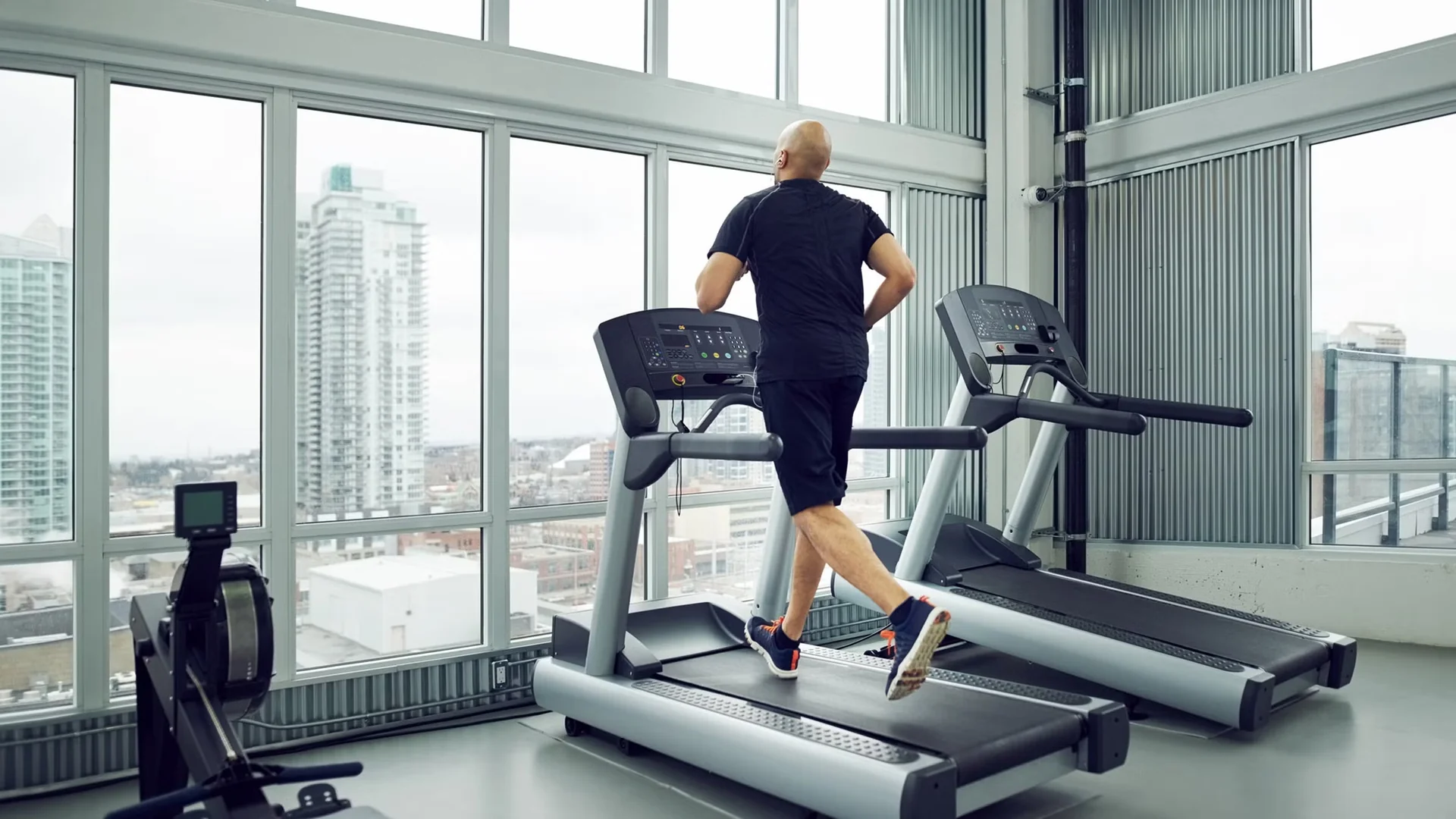 A runner on a treadmill facing floor-to-ceiling windows that overlook a hazy city skyline in a high-rise gym.