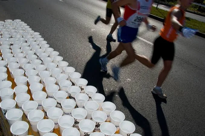 Runners' hands grabbing paper water cups from an aid station table in harsh midday sun.