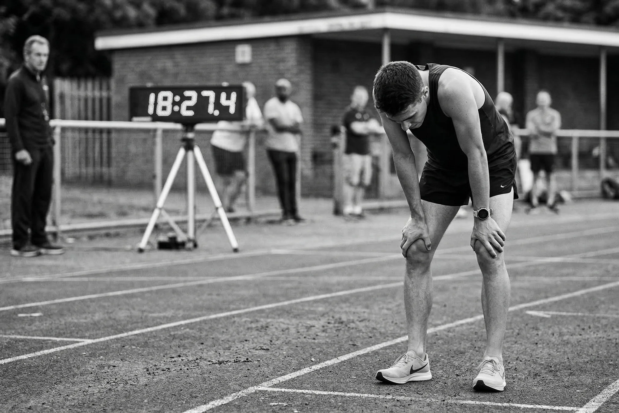 A runner catching their breath with hands on knees after a hard track workout, with a race clock in the background showing their finish time