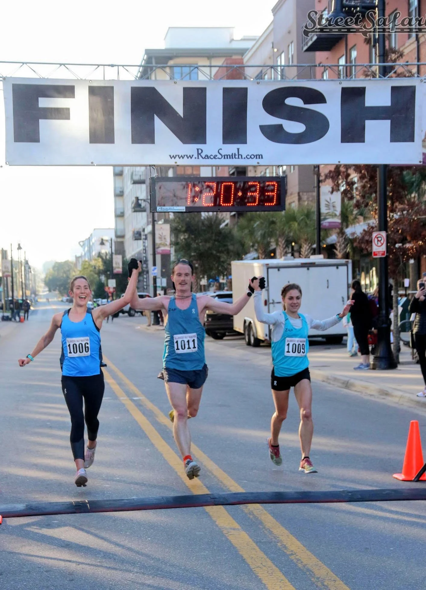 Coach Johnny Crain crossing a half-marathon finish line hand in hand with two athletes at 1:20